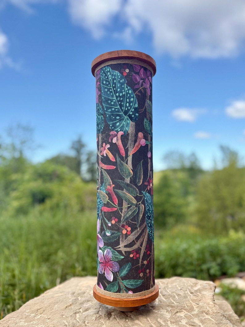 May include: A tall, cylindrical decorative object with a wooden top and base. The body is covered in a dark fabric with a colorful botanical print featuring leaves, flowers, and berries. The object is set against a natural outdoor backdrop.