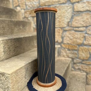 May include: A tall, cylindrical decorative object with a dark blue background and vertical, wavy, light brown lines. The top and bottom rims are made of wood. It sits on a round, woven mat with a dark blue border. The object is placed on stone steps.