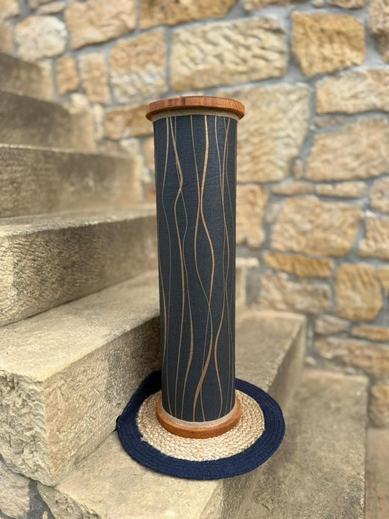 May include: A tall, cylindrical decorative object with a dark blue background and vertical, wavy, light brown lines. The top and bottom rims are made of wood. It sits on a round, woven mat with a dark blue border. The object is placed on stone steps.