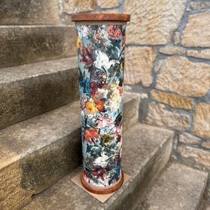 May include: A tall, cylindrical decorative stand with a floral pattern. The stand features a wooden top and base, and the body is covered in a colorful floral print with shades of blue, red, and white. It is placed on a stone staircase.