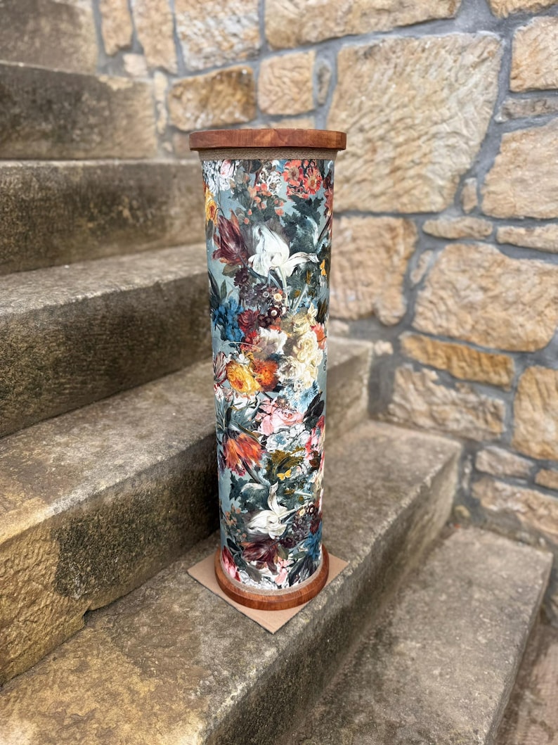May include: A tall, cylindrical decorative stand with a floral pattern. The stand features a wooden top and base, and the body is covered in a colorful floral print with shades of blue, red, and white. It is placed on a stone staircase.