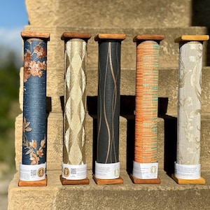 Puede incluir: Se presentan cinco rollos de muestras de papel pintado, cada uno sobre una base de madera y rematado con madera. Los diseños incluyen patrones florales, geométricos y abstractos en azul, dorado, negro, naranja y beige.