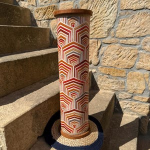 May include: A tall, cylindrical decorative object with a geometric pattern in shades of red, orange, and white. The top and bottom are trimmed with wood. The object is approximately 30 inches tall and is sitting on a round, woven mat.