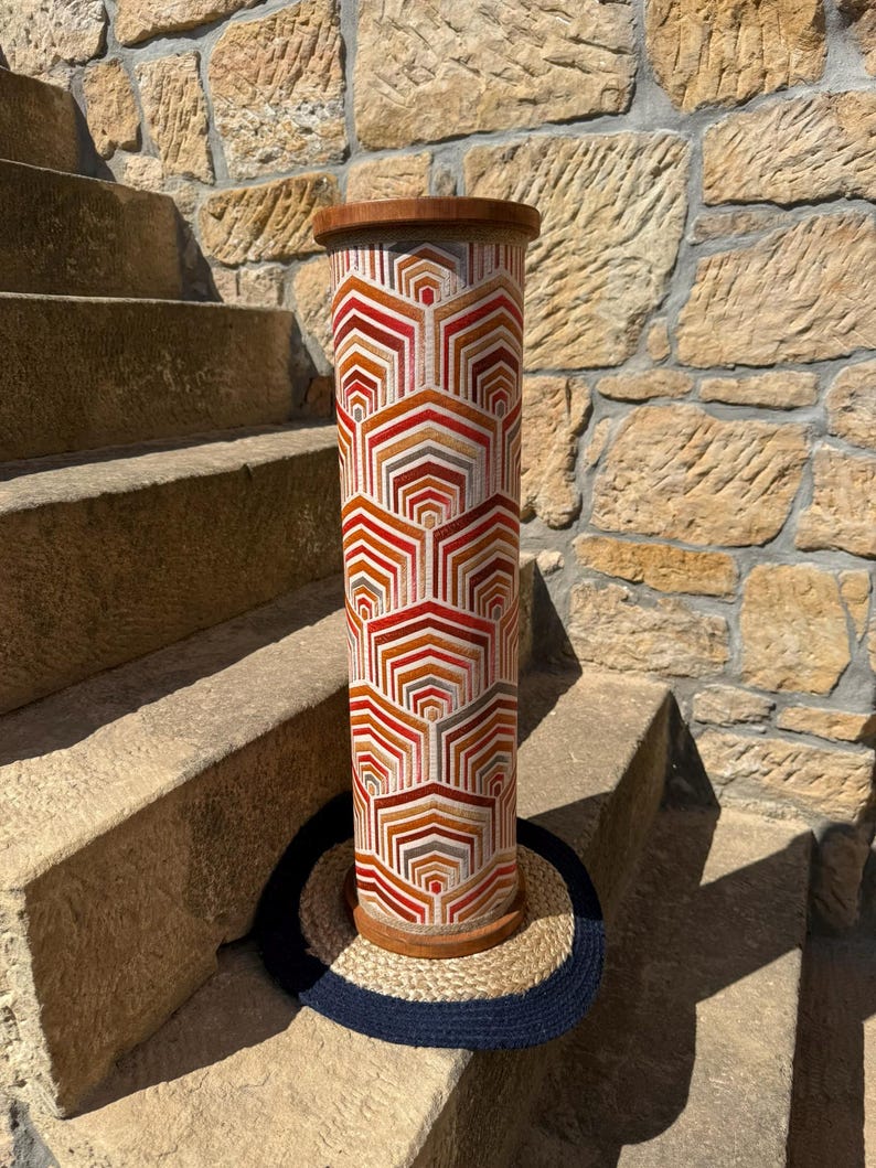 May include: A tall, cylindrical decorative object with a geometric pattern in shades of red, orange, and white. The top and bottom are trimmed with wood. The object is approximately 30 inches tall and is sitting on a round, woven mat.