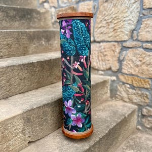May include: A tall, cylindrical decorative item with a wooden top and base. The body is covered in a dark fabric with a vibrant floral pattern featuring teal leaves, pink and purple flowers, and red accents. The item is standing on stone steps.