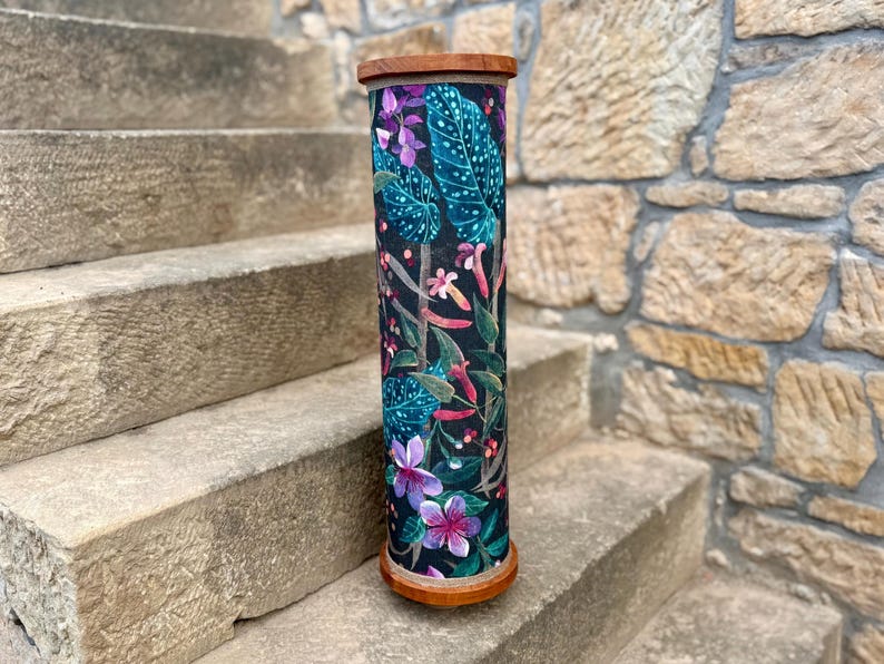 May include: A tall, cylindrical decorative item with a wooden top and base. The body is covered in a dark fabric with a vibrant floral pattern featuring teal leaves, pink and purple flowers, and red accents. The item is standing on stone steps.