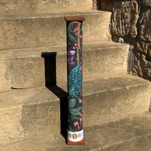 Könnte beinhalten: Ein aufgerolltes, zylindrisches Produkt mit einem botanischen Muster auf dunklem Hintergrund, das grüne, rosa und violette Blätter und Blüten zeigt. Das Produkt wird von einem Holzrahmen gehalten und steht auf einer Steintreppe.