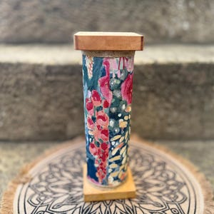 May include: A cylindrical fabric-covered stand featuring a floral pattern in shades of pink, red, blue, and cream. The stand has a square wooden base and a matching square wooden top, with a thin jute-colored trim bordering the fabric edges.