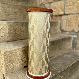 May include: A decorative, cylindrical stand with a geometric pattern. The stand features a light beige fabric with a diamond pattern, and is trimmed with a dark brown wood top and base. The base has a white trim. The stand is approximately 30 inches tall.
