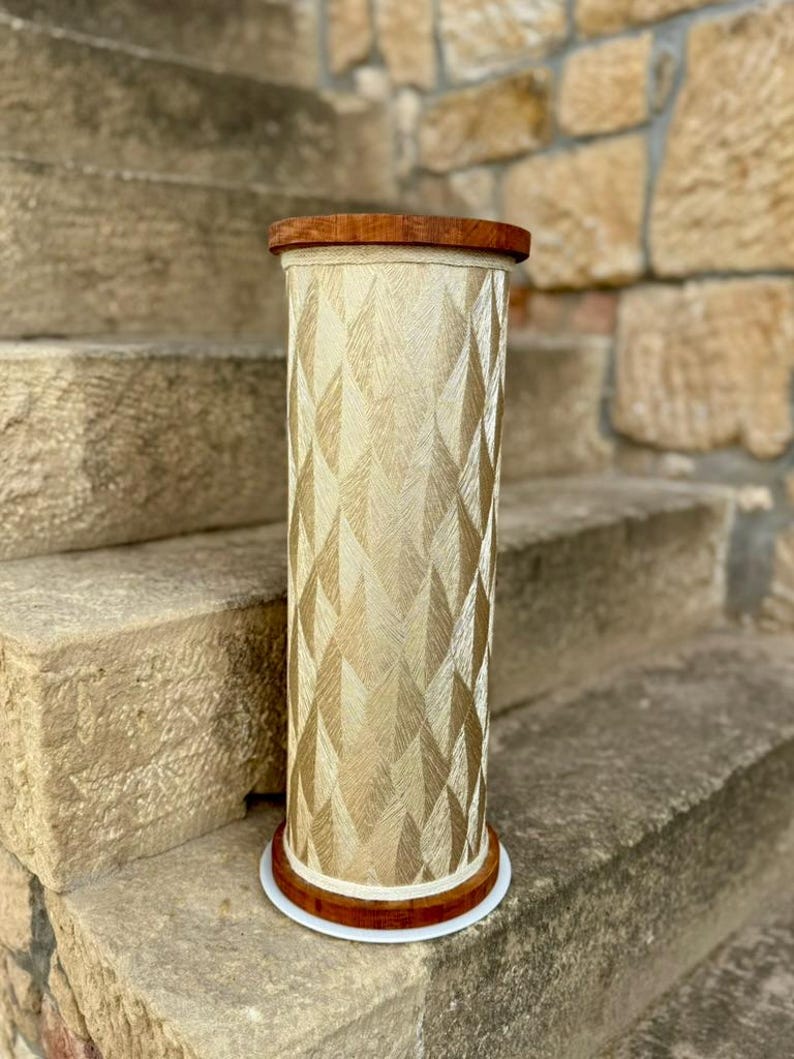 May include: A decorative, cylindrical stand with a geometric pattern. The stand features a light beige fabric with a diamond pattern, and is trimmed with a dark brown wood top and base. The base has a white trim. The stand is approximately 30 inches tall.