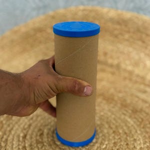 May include: A cylindrical cardboard shipping tube with a light brown finish and a bright blue plastic cap on the top and bottom. The blue cap features embossed circular text that is partially visible, providing a secure closure for the container.