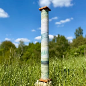 Op de afbeelding: Een hoge, cilindrische keramische sculptuur met een houten boven- en onderkant. De kolom is beschilderd met een mix van blauwe, groene en witte tinten, die op een landschap lijken. Het staat in een veld met hoog, groen gras onder een blauwe hemel.