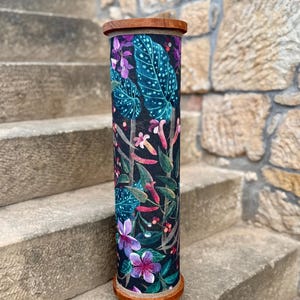 May include: A tall, cylindrical decorative container with a wooden top and base. The container is adorned with a vibrant botanical print featuring large teal leaves, purple flowers, and various other colorful floral elements against a dark background.
