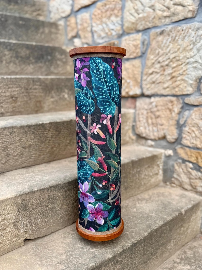May include: A tall, cylindrical decorative container with a wooden top and base. The container is adorned with a vibrant botanical print featuring large teal leaves, purple flowers, and various other colorful floral elements against a dark background.