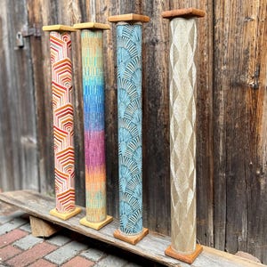 May include: Four decorative wooden pillars with unique designs. The pillars feature geometric, gradient, and fan-like patterns in shades of red, orange, blue, and gold. Each pillar has a square wooden base and top, and they stand on a wooden plank.