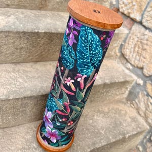 May include: A cylindrical container with a wooden top and base. The body is covered in a fabric with a dark background and a colorful floral pattern featuring teal leaves, purple flowers, and pink accents. The container is resting on stone steps.