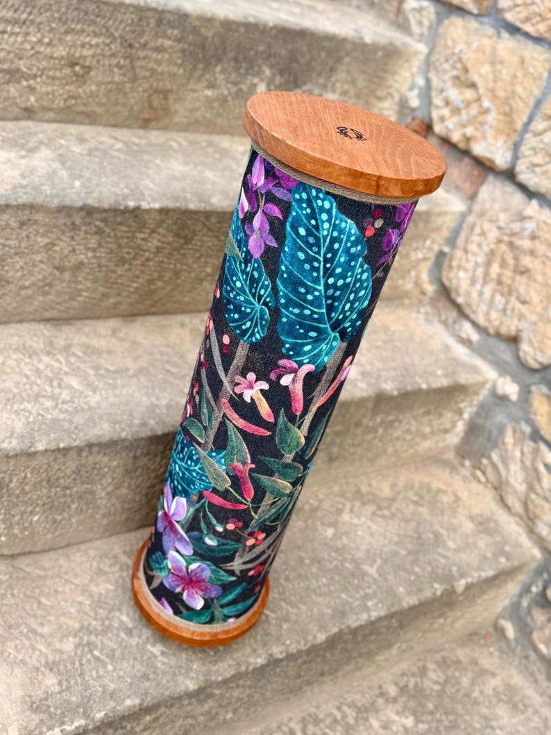 May include: A cylindrical container with a wooden top and base. The body is covered in a fabric with a dark background and a colorful floral pattern featuring teal leaves, purple flowers, and pink accents. The container is resting on stone steps.