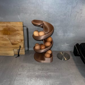 May include: A brown, spiral-shaped egg dispenser rack designed with a gravity-fed track system. The structure features multiple curved tiers that hold several light brown chicken eggs, allowing them to roll down from the top to the base.