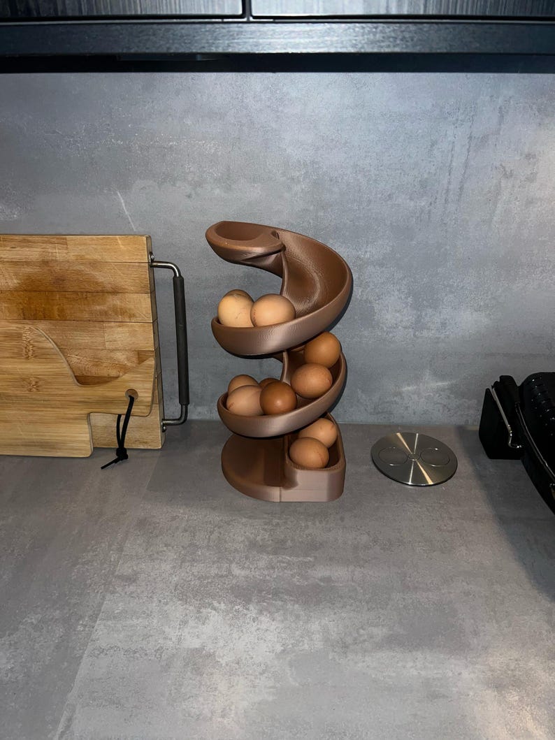 May include: A brown, spiral-shaped egg dispenser rack designed with a gravity-fed track system. The structure features multiple curved tiers that hold several light brown chicken eggs, allowing them to roll down from the top to the base.