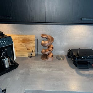 May include: A spiral-shaped egg dispenser made of a matte, light brown material. The structure features a curved, descending track that holds multiple brown eggs in a vertical, stacked arrangement, resting on a circular base on a countertop.