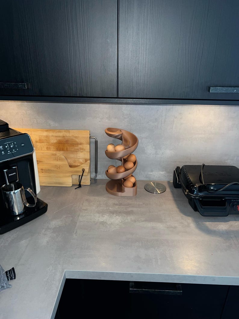 May include: A spiral-shaped egg dispenser made of a matte, light brown material. The structure features a curved, descending track that holds multiple brown eggs in a vertical, stacked arrangement, resting on a circular base on a countertop.
