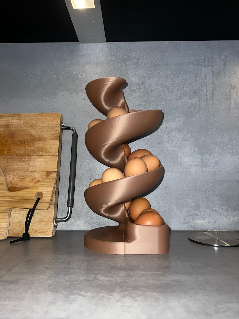 May include: A bronze-colored, spiral-shaped egg dispenser featuring a vertical, winding track design. The structure is constructed with a smooth, layered finish and holds multiple brown eggs that rest along the curved, descending path.
