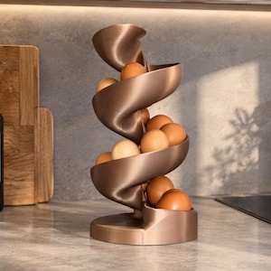 May include: A bronze-colored, spiral-shaped egg dispenser featuring a vertical, winding track design. The structure is crafted with a smooth, metallic finish and holds multiple brown eggs that rest along the curved, descending path to the base.