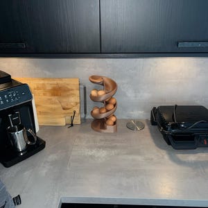 May include: A spiral-shaped egg dispenser made of a matte brown material, designed with a curved track that holds multiple brown eggs in a vertical, descending arrangement, positioned on a gray countertop between kitchen appliances.