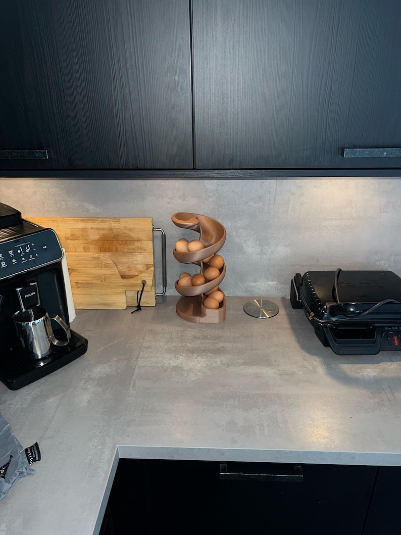 May include: A spiral-shaped egg dispenser made of a matte brown material, designed with a curved track that holds multiple brown eggs in a vertical, descending arrangement, positioned on a gray countertop between kitchen appliances.