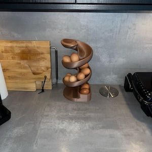 May include: A matte brown spiral egg dispenser with a curved, multi-tiered design holding several light brown eggs. The structure features a continuous track that allows eggs to roll down from the top level to the base for easy access.