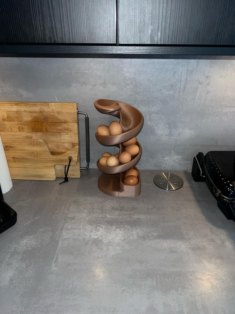 May include: A matte brown spiral egg dispenser with a curved, multi-tiered design holding several light brown eggs. The structure features a continuous track that allows eggs to roll down from the top level to the base for easy access.