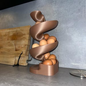 May include: A bronze-colored, 3D-printed spiral egg dispenser featuring a vertical, twisting ramp design. The structure holds multiple brown chicken eggs that rest along the curved track, descending from the top to the base in a continuous flow.