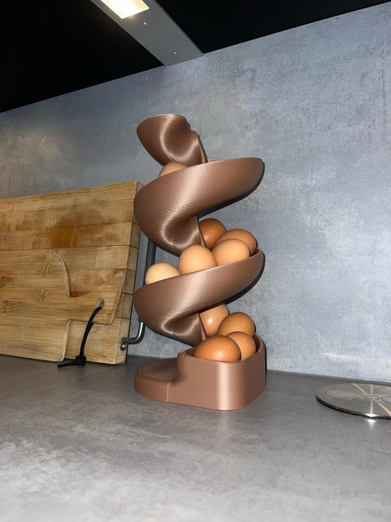 May include: A bronze-colored, 3D-printed spiral egg dispenser featuring a vertical, twisting ramp design. The structure holds multiple brown chicken eggs that rest along the curved track, descending from the top to the base in a continuous flow.