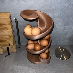 May include: A bronze-colored, spiral-shaped egg dispenser rack with a tiered design. The structure features multiple curved levels that hold several brown chicken eggs, allowing them to roll down the track from the top to the bottom base.