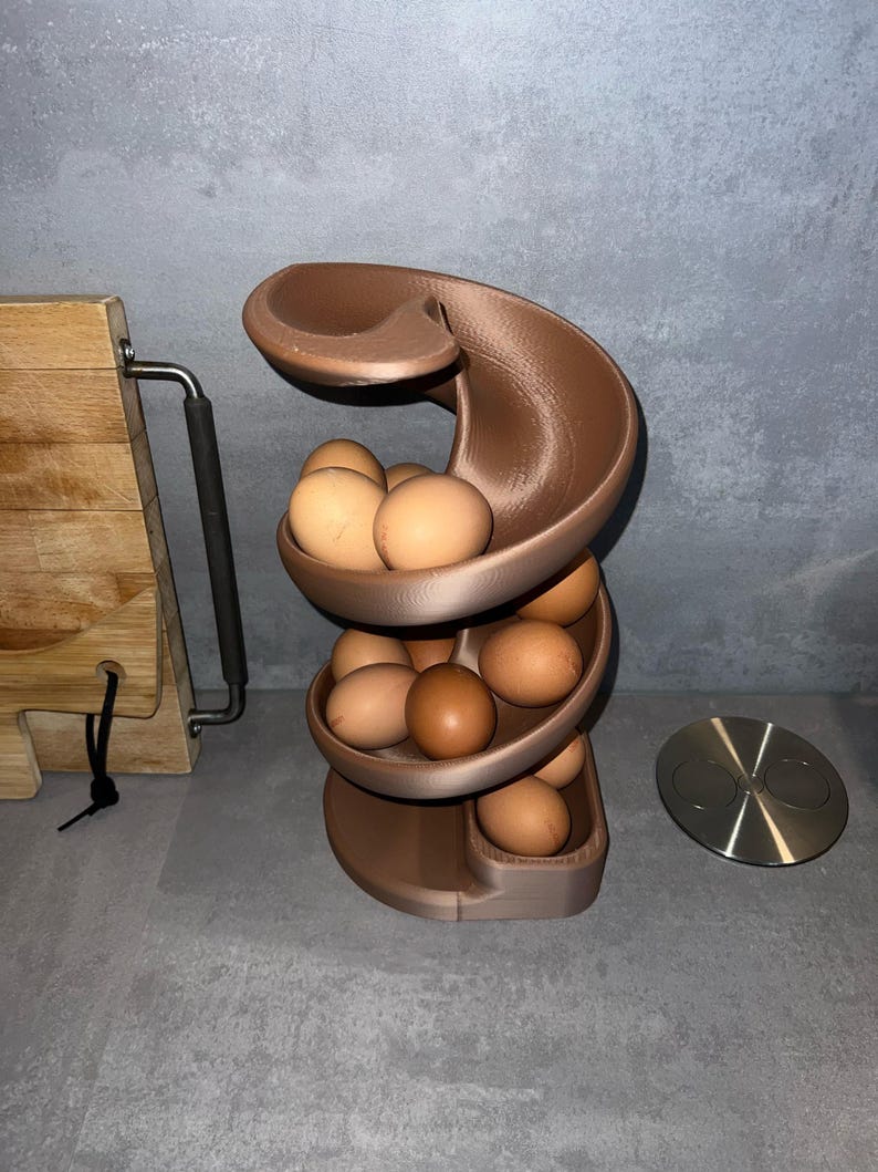 May include: A bronze-colored, spiral-shaped egg dispenser rack with a tiered design. The structure features multiple curved levels that hold several brown chicken eggs, allowing them to roll down the track from the top to the bottom base.