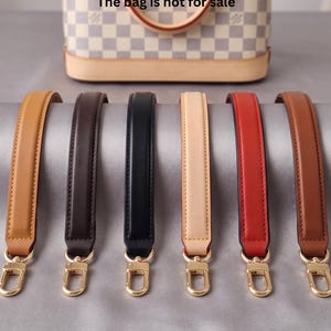 May include: Six leather bag straps in tan, dark brown, black, beige, red, and medium brown, each featuring gold-tone metal lobster clasps at both ends. A checkered handbag is visible in the background with the text The bag is not for sale.
