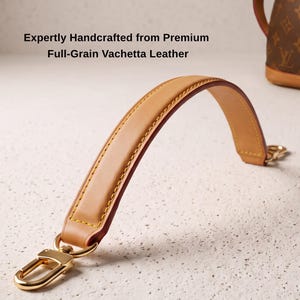 Könnte beinhalten: Ein beigefarbener Handtaschenriemen aus Vachetta-Leder mit goldfarbenen Karabinerverschlüssen aus Metall an beiden Enden. Der Riemen hat gelbe Nähte an den Kanten. Der Text lautet: Expertly Handcrafted from Premium Full-Grain Vachetta Leather.