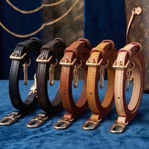 May include: Five adjustable handbag straps in a row, ranging in color from black to dark brown, medium brown, tan, and light beige. Each strap features gold-tone metal buckles and lobster claw clasps for attachment, with visible stitching along the edges.