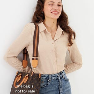 Op de afbeelding: Bruine handtas met een patroon en een bruine en bruin/beige schouderriem. De tas heeft een handvat aan de bovenkant en de tekst "The bag is not for sale". De vrouw draagt een beige overhemd en een blauwe jeans.
