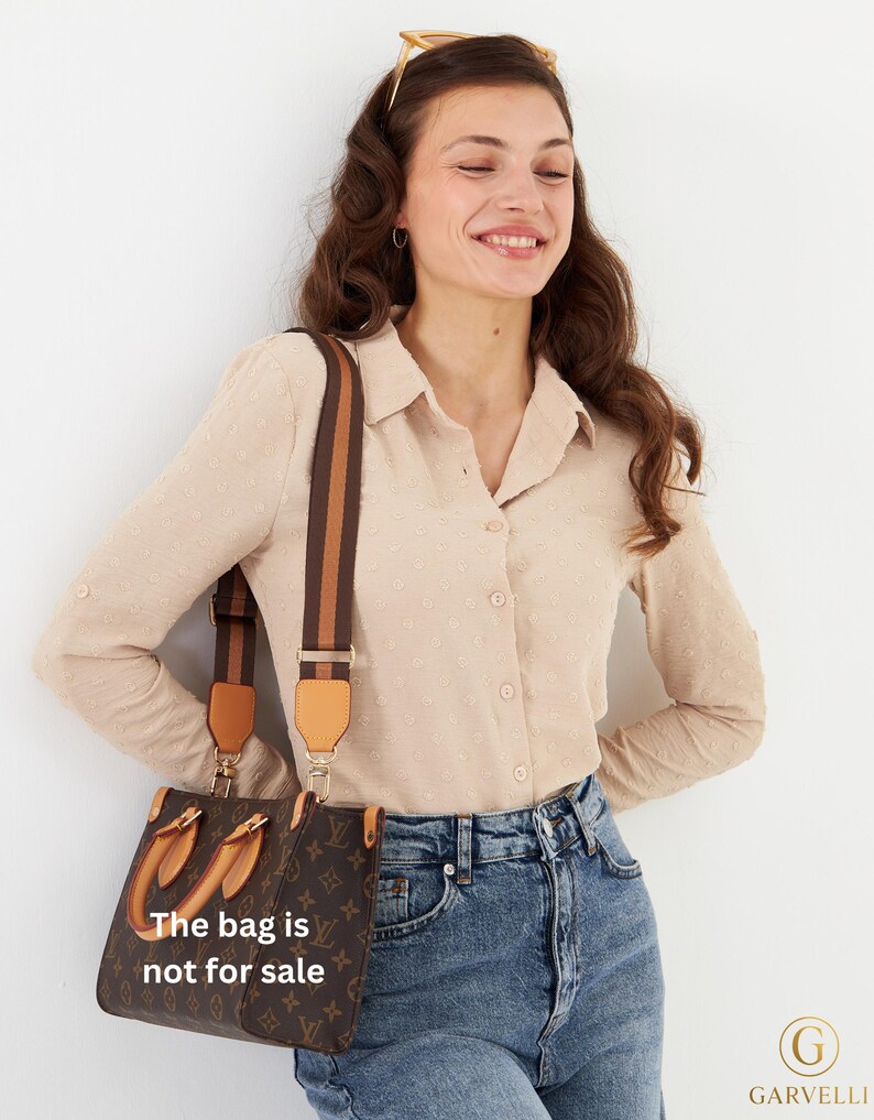 Op de afbeelding: Bruine handtas met een patroon en een bruine en bruin/beige schouderriem. De tas heeft een handvat aan de bovenkant en de tekst "The bag is not for sale". De vrouw draagt een beige overhemd en een blauwe jeans.