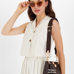 May include: A woman wearing light brown sunglasses and a cream-colored outfit, holding the sunglasses. A brown monogrammed handbag with a brown strap hangs from her shoulder. The text on the bag reads, "The bag is not for sale."
