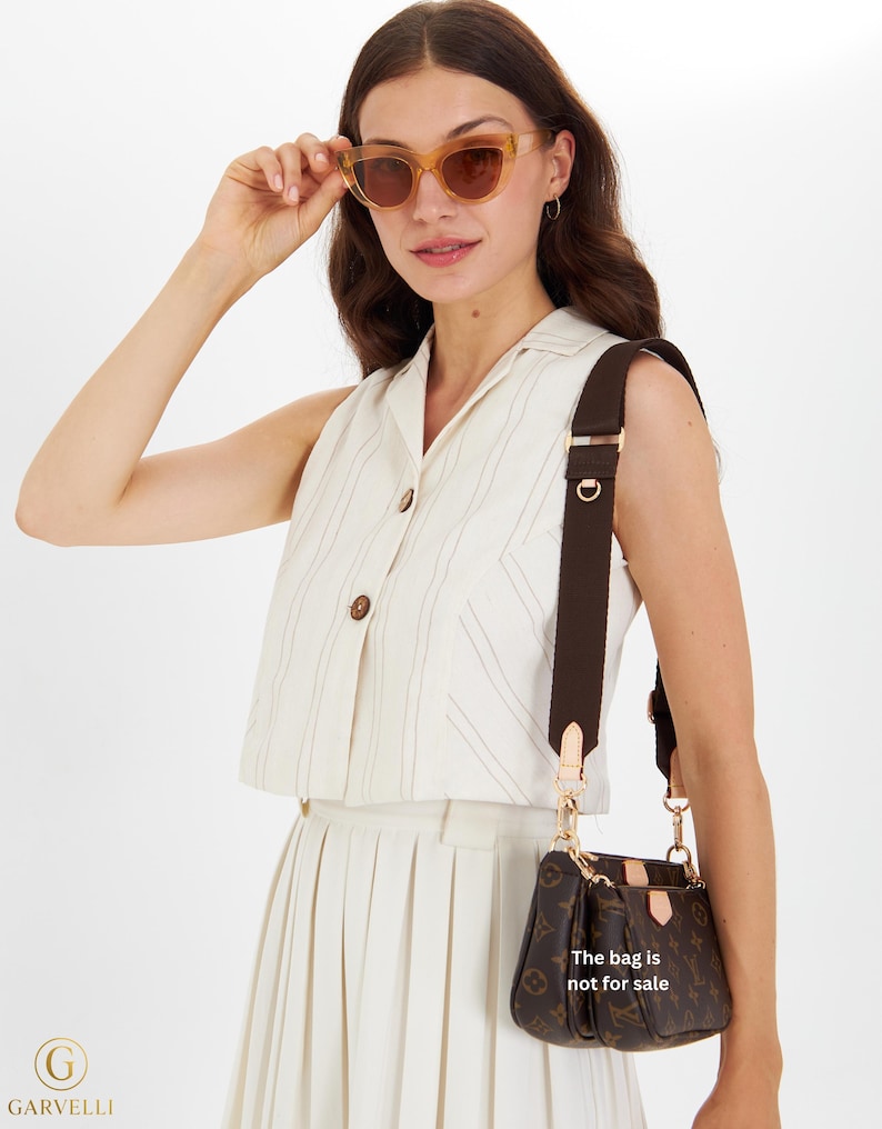 May include: A woman wearing light brown sunglasses and a cream-colored outfit, holding the sunglasses. A brown monogrammed handbag with a brown strap hangs from her shoulder. The text on the bag reads, "The bag is not for sale."