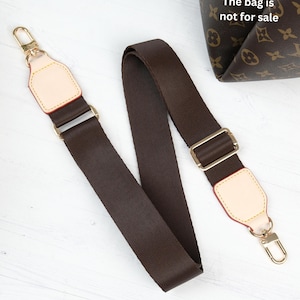 Op de afbeelding: Een bruine tasriem met goudkleurige hardware en beige lederen accenten. De riem is gemaakt van een duurzaam, geweven materiaal. De tas op de achtergrond heeft een bruin en beige patroon. De tekst op de tas luidt "The bag is not for sale."