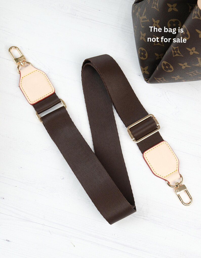 Op de afbeelding: Een bruine tasriem met goudkleurige hardware en beige lederen accenten. De riem is gemaakt van een duurzaam, geweven materiaal. De tas op de achtergrond heeft een bruin en beige patroon. De tekst op de tas luidt "The bag is not for sale."