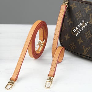 Puede incluir: Correa de bolso de cuero color canela con herrajes dorados y bordes rojos. La correa se muestra enrollada y extendida, con un bolso a juego en el fondo. El bolso tiene un patr&oacute;n de monograma marr&oacute;n. El texto en el bolso dice "La bolsa no est&aacute; a la venta."