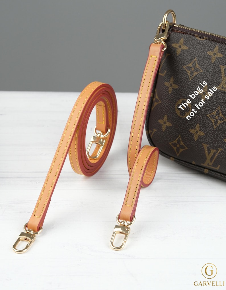 Puede incluir: Correa de bolso de cuero color canela con herrajes dorados y bordes rojos. La correa se muestra enrollada y extendida, con un bolso a juego en el fondo. El bolso tiene un patr&oacute;n de monograma marr&oacute;n. El texto en el bolso dice "La bolsa no est&aacute; a la venta."