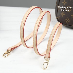 Puede incluir: Una correa de bolso de cuero beige claro con bordes rojos y cierres met&aacute;licos dorados. La correa est&aacute; cosida con un hilo en contraste. El texto "The bag is not for sale" es visible en el fondo.