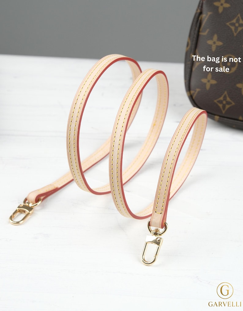 Puede incluir: Una correa de bolso de cuero beige claro con bordes rojos y cierres met&aacute;licos dorados. La correa est&aacute; cosida con un hilo en contraste. El texto "The bag is not for sale" es visible en el fondo.
