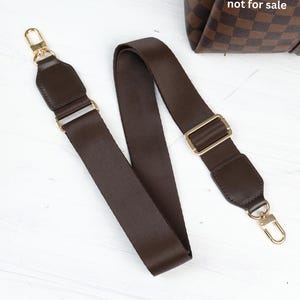 Op de afbeelding: Een bruine tasriem met goudkleurige hardware. De riem is gemaakt van een geweven materiaal en heeft leren accenten aan elk uiteinde. De tekst "The bag is not for sale" is zichtbaar op de achtergrond.
