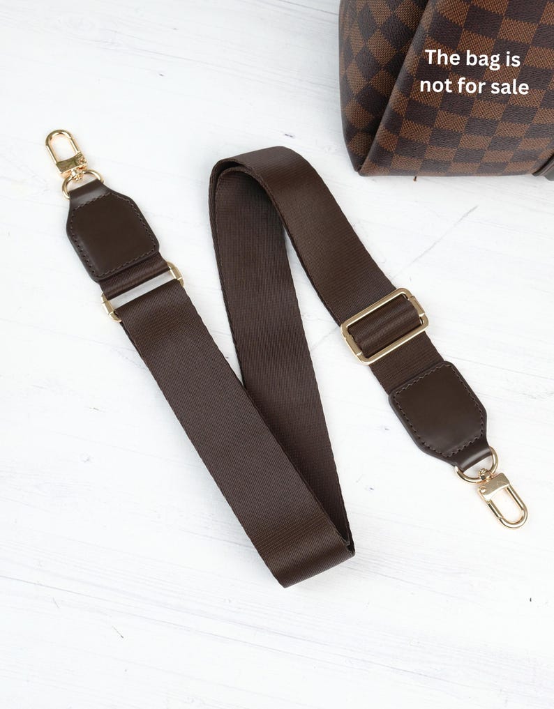 Op de afbeelding: Een bruine tasriem met goudkleurige hardware. De riem is gemaakt van een geweven materiaal en heeft leren accenten aan elk uiteinde. De tekst "The bag is not for sale" is zichtbaar op de achtergrond.